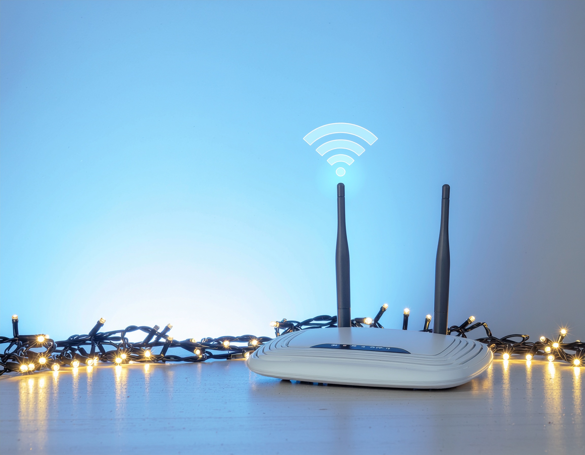 A white wireless router with two black antennas emits a glowing Wi-Fi signal icon against a soft blue background. The router is surrounded by festive string lights, symbolizing strong wireless connectivity and the role of D-Link QoS settings in maintaining seamless internet performance.