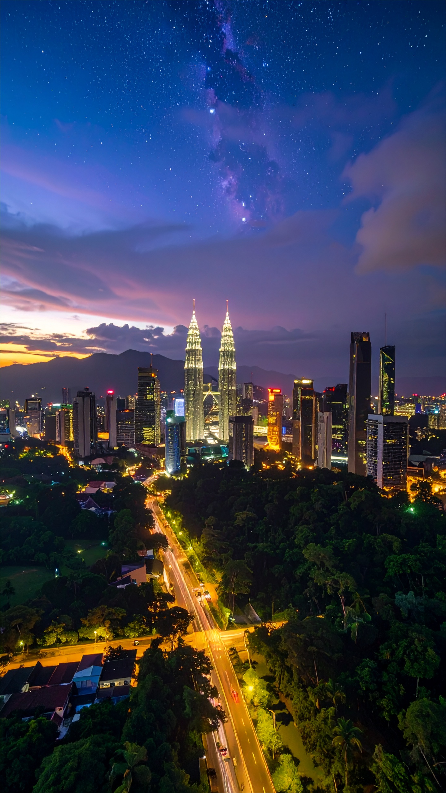 Nighttime cityscape of Kuala Lumpur featuring the illuminated Petronas Twin Towers surrounded by modern skyscrapers, with vibrant streetlights and the Milky Way visible in the starry sky above.